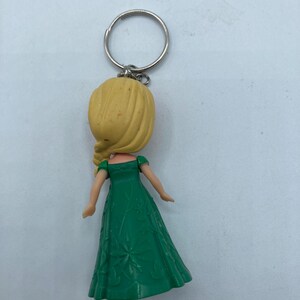 May include: A keychain featuring a doll with a yellow head and blonde hair, wearing a green dress. The dress has a floral pattern. The keychain ring is silver.