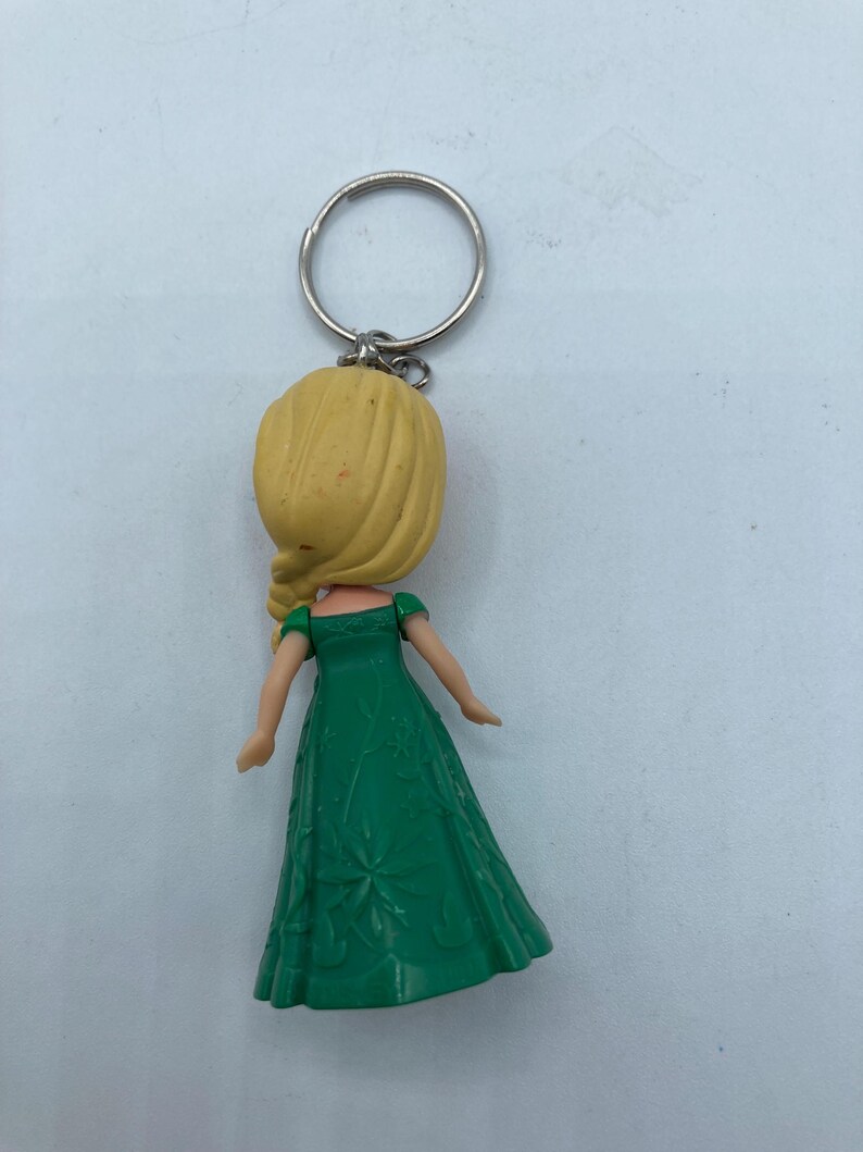 May include: A keychain featuring a doll with a yellow head and blonde hair, wearing a green dress. The dress has a floral pattern. The keychain ring is silver.