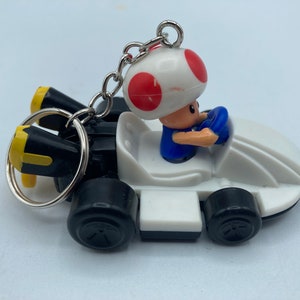 May include: A keychain featuring a white race car with black wheels and yellow accents. A small figure of Toad, wearing a blue shirt and white mushroom cap with red spots, is driving the car. The keychain has a silver ring.