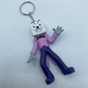 May include: A novelty keychain featuring a cartoon character with a dice-shaped head, wearing a pink jacket, purple pants, and a black bow tie. The character has a wide, toothy grin and gray gloves. The keychain is attached to a silver ring.