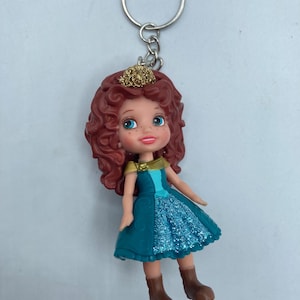 May include: A keychain featuring a doll with red curly hair, blue eyes, and a gold crown. The doll wears a teal dress with a glittery blue skirt and brown boots. The keychain has a silver ring.
