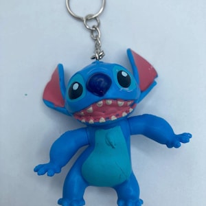 K&ouml;nnte beinhalten: Ein blauer Stitch-Schl&uuml;sselanh&auml;nger mit den charakteristischen gro&szlig;en Augen, rosa Ohren und offenem Mund des Charakters mit sichtbaren Z&auml;hnen. Der Schl&uuml;sselanh&auml;nger hat einen silbernen Metallring zum Befestigen an Schl&uuml;sseln oder Taschen. Die Figur ist leuchtend blau mit einem helleren blauen Bauch.