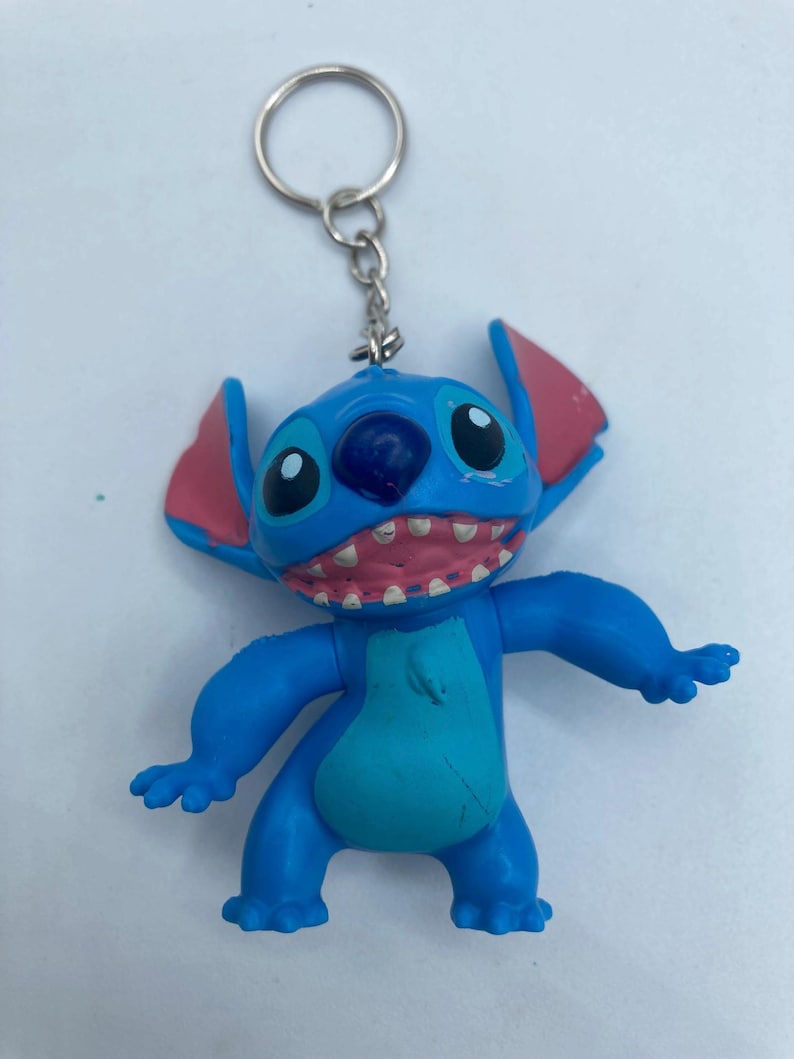 K&ouml;nnte beinhalten: Ein blauer Stitch-Schl&uuml;sselanh&auml;nger mit den charakteristischen gro&szlig;en Augen, rosa Ohren und offenem Mund des Charakters mit sichtbaren Z&auml;hnen. Der Schl&uuml;sselanh&auml;nger hat einen silbernen Metallring zum Befestigen an Schl&uuml;sseln oder Taschen. Die Figur ist leuchtend blau mit einem helleren blauen Bauch.