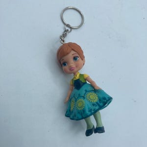 May include: A small keychain featuring a doll with red hair, blue eyes, and a teal dress with yellow sunflower designs. The doll has a yellow shirt and green vest. The keychain is made of metal.