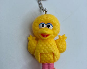 May include: A yellow Big Bird keychain featuring the Sesame Street character. The keychain has a silver metal ring and chain. Big Bird has blue eyes, an orange beak, and orange feet. The keychain is made of plastic.