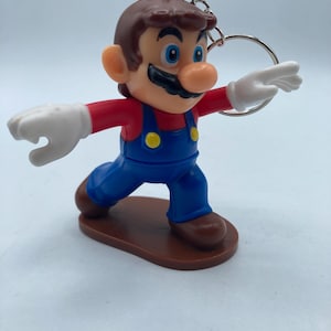 May include: A keychain featuring a figurine of a cartoon character in a red shirt, blue overalls, and brown shoes. The character has a large black mustache, brown hair, and white gloves. The figure is attached to a silver keyring.
