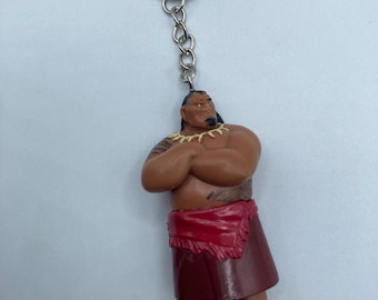May include: A keychain featuring a figurine of a muscular character with crossed arms. The figure has brown skin, a red skirt, and a necklace. The keychain has a silver ring and chain.