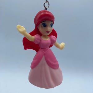 May include: Ariel keychain featuring a miniature figurine of the Disney princess. The figure has red hair, blue eyes, and is wearing a pink dress. The keychain is attached to a silver chain.