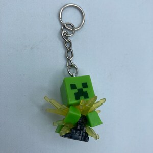 May include: A green Minecraft Creeper keychain with a yellow glow effect. The keychain is attached to a silver metal chain and ring.