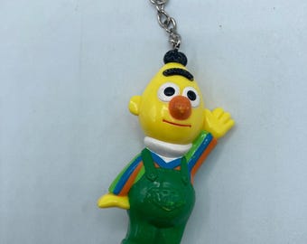 May include: A yellow Bert keychain from Sesame Street. Bert is wearing green overalls with a white collar and a black hat. The keychain has a silver ring and chain.