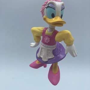May include: A white duck figurine with pink hair, a yellow and pink dress, and pink shoes. The duck is wearing a white apron with a floral pattern. The figurine is hanging from a silver chain.