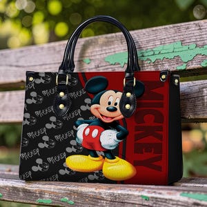 May include: Black tote bag featuring Mickey Mouse. The bag has black handles and gold-colored hardware. The front of the bag has a red panel with the word "Mickey" in large letters. The left side of the bag has a black background with Mickey Mouse silhouettes.