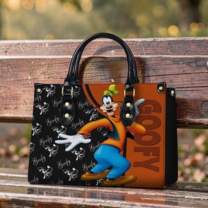 May include: A black and brown handbag featuring a cartoon image of Goofy. The bag has black handles and gold-colored hardware. The left side of the bag has a black background with a white Goofy pattern, while the right side is brown with the word "GOOFY".