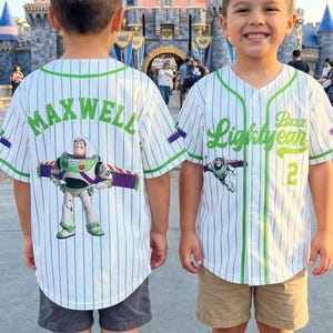 K&ouml;nnte beinhalten: Zwei Kinder tragen wei&szlig;e Baseballtrikots mit gr&uuml;nen Nadelstreifen und Buzz Lightyear-Grafiken. Auf einem Trikot steht "Maxwell" auf dem R&uuml;cken, auf dem anderen "Buzz Lightyear" und die Nummer 2. Beide tragen Shorts und Turnschuhe.