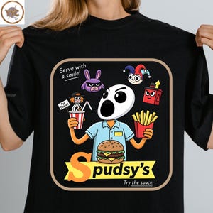 May include: A black t-shirt featuring a graphic of a character with a white oval head, wearing a light blue uniform, holding a drink and fries with a burger in front. Text reads Serve with a smile!, For Caine!, Spudsy's, and Try the sauce.