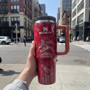 Peut inclure: Mug de voyage Stanley rouge avec un motif argenté de Spider-Man. Le mug a un couvercle noir, une poignée pêche et le logo Stanley. Le design comprend les mots "Amazing Spider-Man".