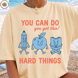 May include: A cream-colored t-shirt featuring a graphic print with the text "YOU CAN DO you got this! HARD THINGS" in orange and red. Below the text are three light blue cartoon characters: a pencil, an apple, and an open book with faces.