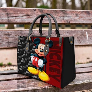 May include: Black tote bag featuring Mickey Mouse. The bag has a red panel with the word "Mickey" in large letters. The other side of the bag has a pattern of Mickey Mouse faces and the word "Mickey". The bag has black handles and gold-colored hardware.