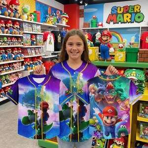May include: A young person holds up three Super Mario baseball jerseys. The jerseys feature colorful graphics of Mario, Yoshi, and other characters against a space-themed background. The store's sign reads "Super Mario".