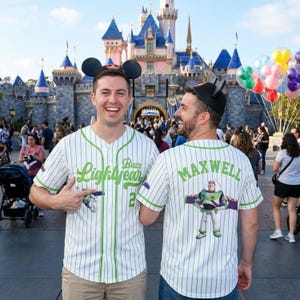K&ouml;nnte beinhalten: Zwei Personen tragen wei&szlig;e Baseball-Trikots mit gr&uuml;nen Nadelstreifen und den Texten "Buzz Lightyear" und "Maxwell". Die Trikots zeigen ein Cartoon-Bild von Buzz Lightyear. Sie tragen Mickey Mouse Ohren vor einem Schloss.