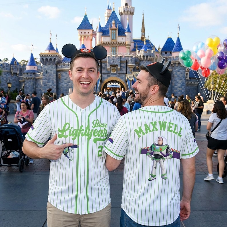K&ouml;nnte beinhalten: Zwei Personen tragen wei&szlig;e Baseball-Trikots mit gr&uuml;nen Nadelstreifen und den Texten "Buzz Lightyear" und "Maxwell". Die Trikots zeigen ein Cartoon-Bild von Buzz Lightyear. Sie tragen Mickey Mouse Ohren vor einem Schloss.