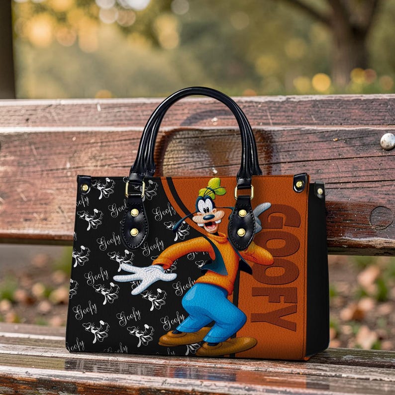 May include: A black and brown handbag featuring a cartoon image of Goofy. The bag has black handles and gold-colored hardware. The left side of the bag has a black background with a white Goofy pattern, while the right side is brown with the word "GOOFY".