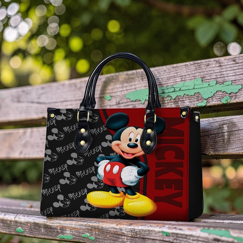 May include: Black tote bag featuring Mickey Mouse. The bag has black handles and gold-colored hardware. The front of the bag has a red panel with the word "Mickey" in large letters. The left side of the bag has a black background with Mickey Mouse silhouettes.