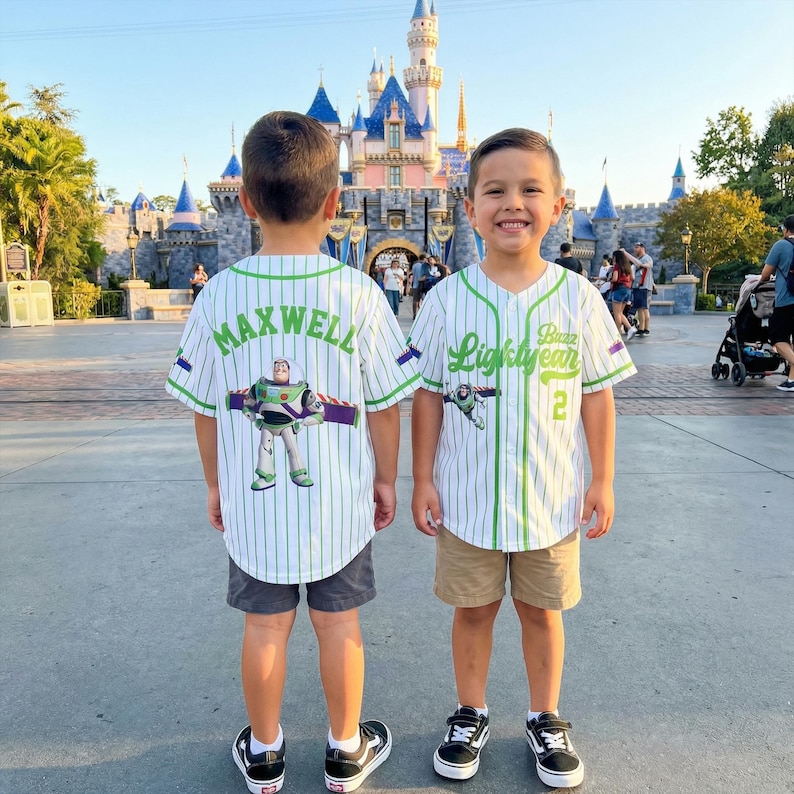K&ouml;nnte beinhalten: Zwei Kinder tragen wei&szlig;e Baseballtrikots mit gr&uuml;nen Nadelstreifen und Buzz Lightyear-Grafiken. Auf einem Trikot steht "Maxwell" auf dem R&uuml;cken, auf dem anderen "Buzz Lightyear" und die Nummer 2. Beide tragen Shorts und Turnschuhe.