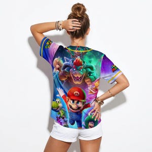May include: A short-sleeved t-shirt featuring a vibrant graphic of characters from the Super Mario video game series. The design includes Mario, Bowser, Princess Peach, and other recognizable figures, set against a colorful, space-themed background.