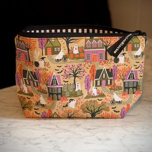 May include: A zippered pouch featuring a tan background with a repeating pattern of houses, trees, pumpkins, and ghosts on bicycles. The fabric has a black zipper closure with a black fabric tag labeled Stolen Minutes and a black and white striped lining.