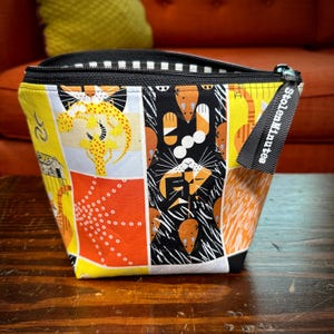 Könnte beinhalten: Eine schwarze Reißverschlusstasche mit einem bunten Patchwork-Design mit Katzen, Mäusen und geometrischen Mustern. Die Tasche hat einen schwarzen Reißverschluss mit dem aufgedruckten Text "Stolen Minutes".