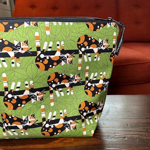 Puede incluir: Una bolsa de tela verde con cremallera negra y tirador negro. La bolsa presenta un estampado repetido de gatos negros, naranjas y blancos sobre un fondo de hojas verdes. La bolsa est&aacute; sobre una superficie de madera oscura.