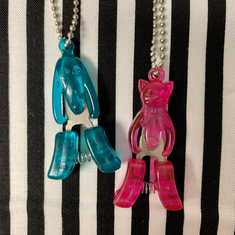 K&ouml;nnte beinhalten: Zwei Tier-f&ouml;rmige Halsketten aus Plastik. Die Halskette links ist blau und hat die Form eines Hundes. Die Halskette rechts ist pink und hat die Form einer Katze.