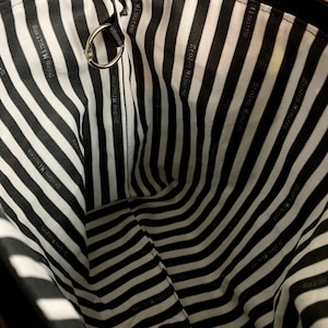 Puede incluir: Vista interior de una bolsa negra con cremallera y forro a rayas blancas y negras. El forro presenta el texto "Stolen Minutes" en un patr&oacute;n repetido. Un anillo de metal plateado est&aacute; sujeto al borde superior.