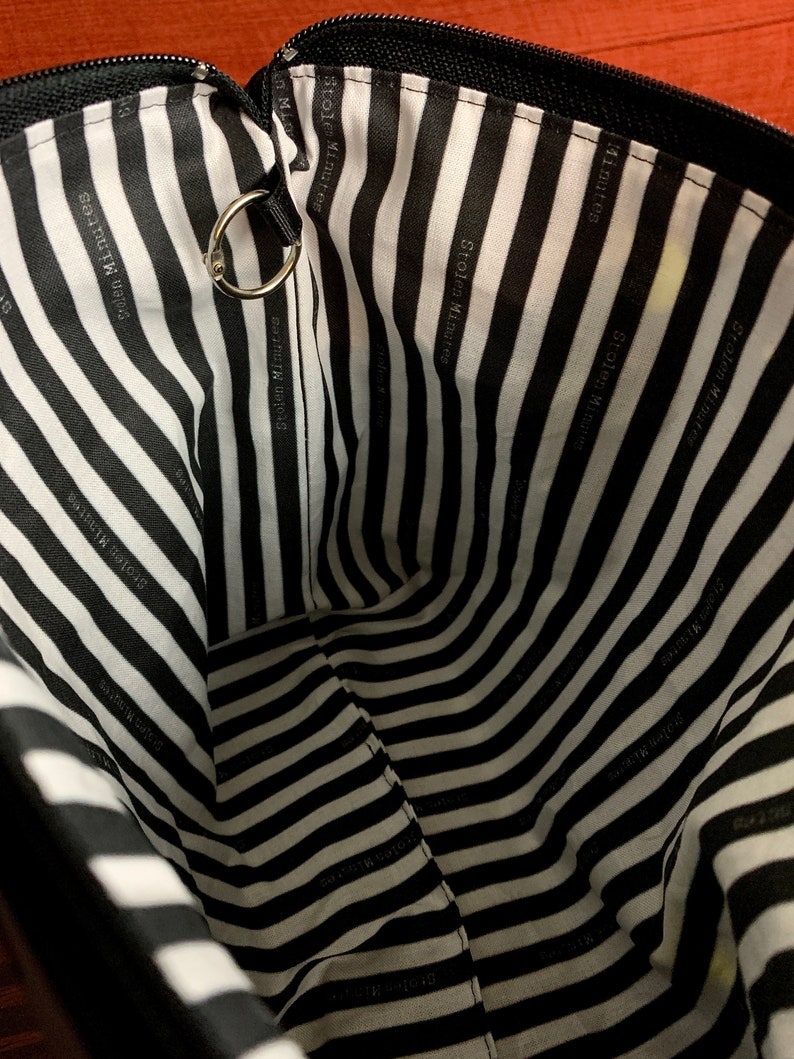 Puede incluir: Vista interior de una bolsa negra con cremallera y forro a rayas blancas y negras. El forro presenta el texto "Stolen Minutes" en un patr&oacute;n repetido. Un anillo de metal plateado est&aacute; sujeto al borde superior.