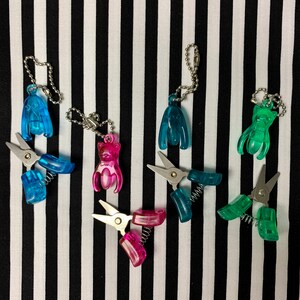 K&ouml;nnte beinhalten: F&uuml;nf bunte Schl&uuml;sselanh&auml;nger mit kleinen Scheren. Die Schl&uuml;sselanh&auml;nger zeigen verschiedene Tierformen, darunter eine Katze, einen Hund und einen Vogel. Die Scheren sind silberfarben und haben einen Federmechanismus.