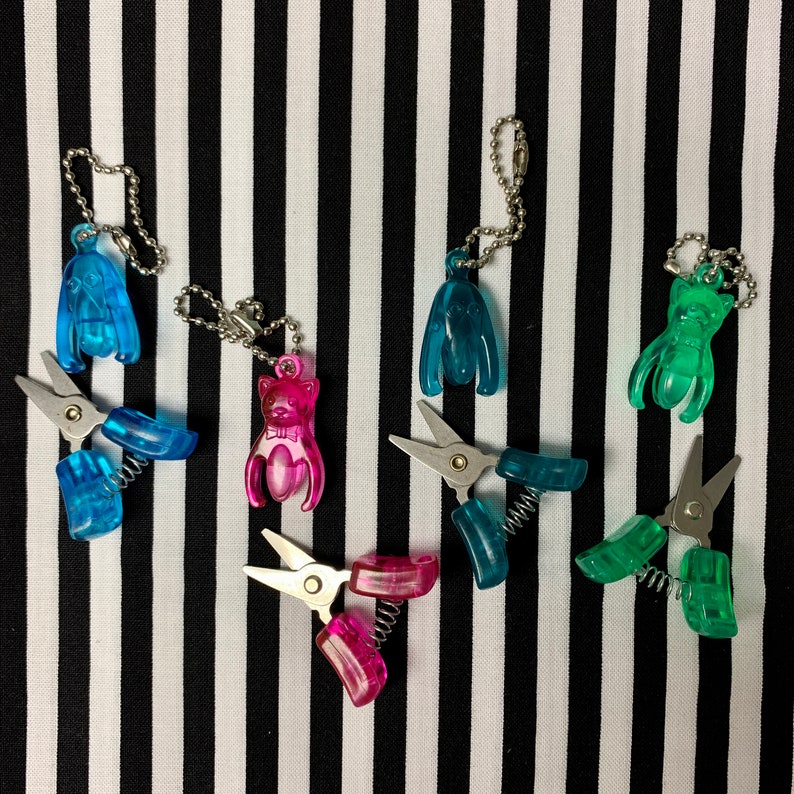 K&ouml;nnte beinhalten: F&uuml;nf bunte Schl&uuml;sselanh&auml;nger mit kleinen Scheren. Die Schl&uuml;sselanh&auml;nger zeigen verschiedene Tierformen, darunter eine Katze, einen Hund und einen Vogel. Die Scheren sind silberfarben und haben einen Federmechanismus.