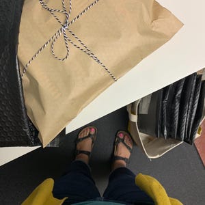 Puede incluir: Un paquete envuelto en papel marr&oacute;n y atado con hilo blanco y negro. El paquete est&aacute; colocado sobre una superficie blanca junto a un sobre de burbujas negro. La persona lleva sandalias y un c&aacute;rdigan amarillo.