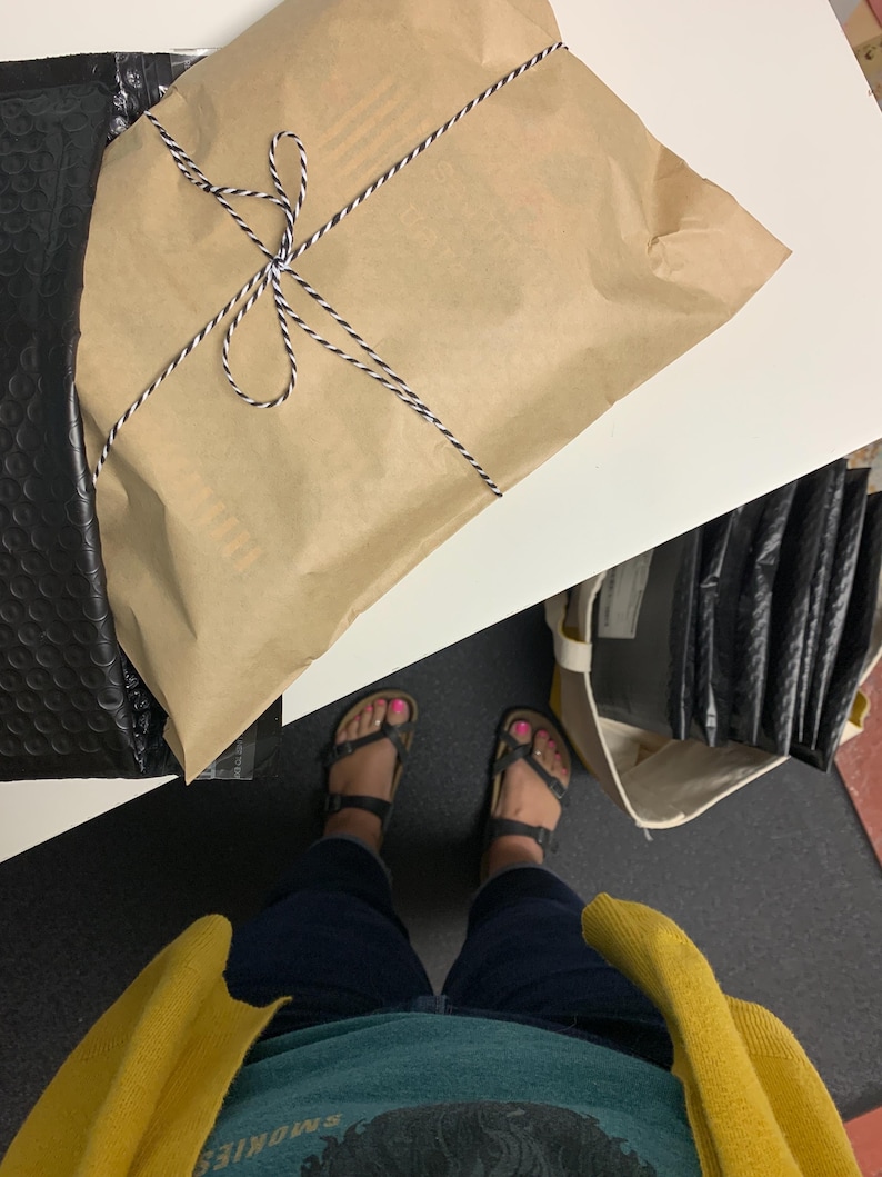 Puede incluir: Un paquete envuelto en papel marr&oacute;n y atado con hilo blanco y negro. El paquete est&aacute; colocado sobre una superficie blanca junto a un sobre de burbujas negro. La persona lleva sandalias y un c&aacute;rdigan amarillo.
