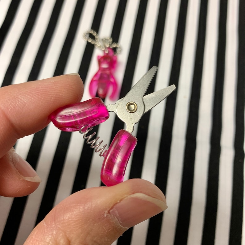 K&ouml;nnte beinhalten: Eine kleine rosa Schere mit einem Federmechanismus. Die Schere hat eine silberne Klinge und wird in einer Hand gehalten.