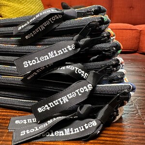 Puede incluir: Una pila de bolsas negras con cremallera y textura tejida. Cada bolsa tiene una etiqueta de cinta en blanco y negro que dice "StolenMinutes" en texto blanco. Las bolsas est&aacute;n sobre una superficie de madera.