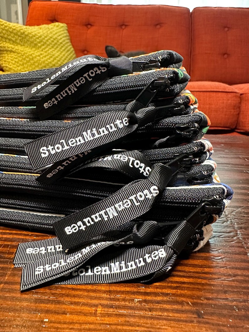 Puede incluir: Una pila de bolsas negras con cremallera y textura tejida. Cada bolsa tiene una etiqueta de cinta en blanco y negro que dice "StolenMinutes" en texto blanco. Las bolsas est&aacute;n sobre una superficie de madera.