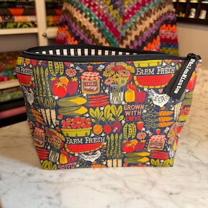 Op de afbeelding: Een etui met rits op een donkerblauwe achtergrond met een kleurrijke boerderijprint van groenten, fruit, honingpotten en kippen. De tekst "FARM FRESH" en "GROWN WITH LOVE" is overal gedrukt. Een zwart label bevat de tekst "StolenMinutes".