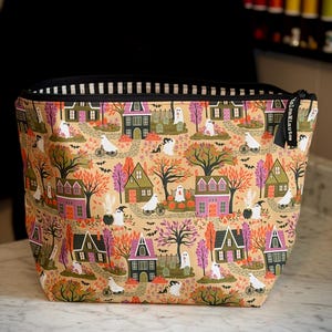 May include: A zippered pouch featuring a tan background with a repeating pattern of colorful houses, trees, ghosts, and pumpkins. The fabric print includes small black bats and ghosts riding bicycles. The pouch has a black zipper and a black tag.