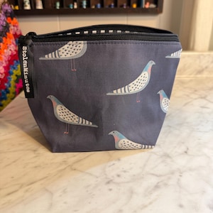 Könnte beinhalten: Eine dunkelgraue Reißverschlusstasche mit einem sich wiederholenden Muster aus stilisierten Vögeln mit weißen, blauen und rosa Details. Ein schwarzes Stoffetikett an der Seite des Reißverschlusses ist mit dem weißen Text StolenMinutes bedruckt. Das Innenfutter ist schwarz-weiß.