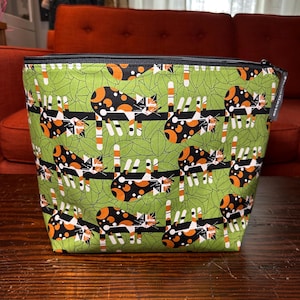 May include: A square, zippered bag with a green background and a repeating pattern of calico cats. The cats are black, orange, and white, and are positioned on a black fence. The bag has a black zipper and a black lining.