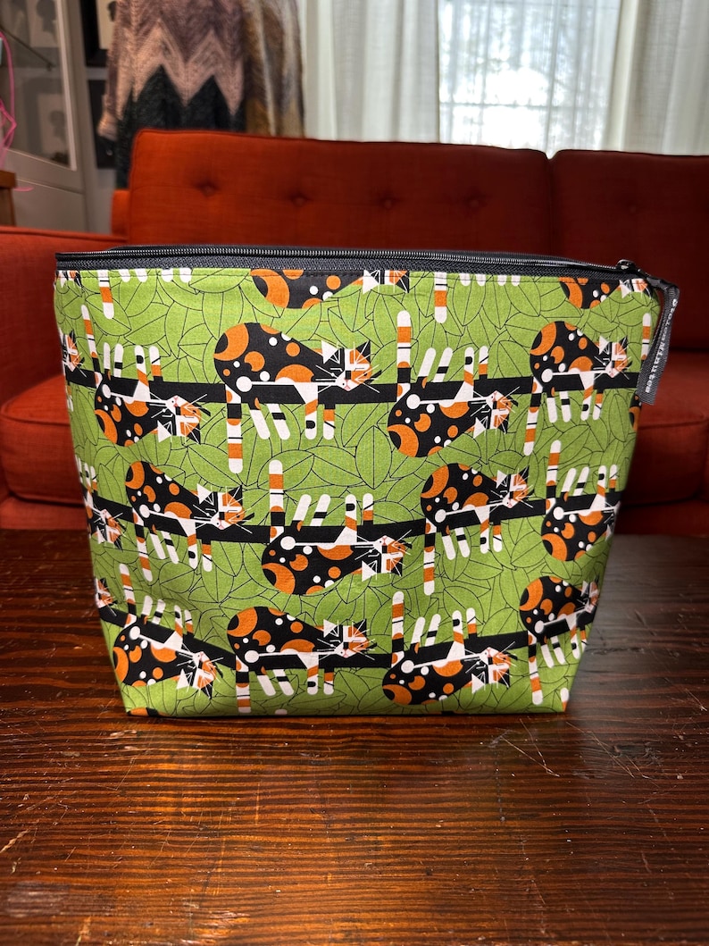 Puede incluir: Una bolsa cuadrada con cremallera, fondo verde y un patr&oacute;n repetido de gatos calic&oacute;. Los gatos son negros, naranjas y blancos, y est&aacute;n colocados sobre una valla negra. La bolsa tiene una cremallera negra y un forro negro.