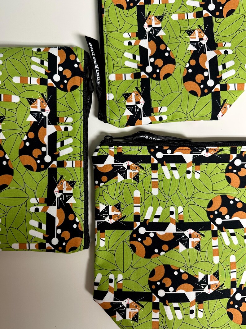 Puede incluir: Tres estuches con cremallera con un patr&oacute;n repetido de gatos estilizados en negro, naranja y blanco sobre un fondo de hojas verdes. Los estuches var&iacute;an en tama&ntilde;o y est&aacute;n dispuestos sobre una superficie blanca. El dise&ntilde;o incluye el texto "stolenMinute".