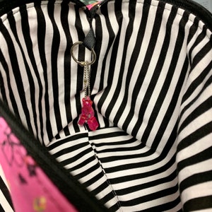 K&ouml;nnte beinhalten: Das Innere einer schwarz-wei&szlig; gestreiften Tasche mit einem rosa Anh&auml;nger an einer silbernen Kette.