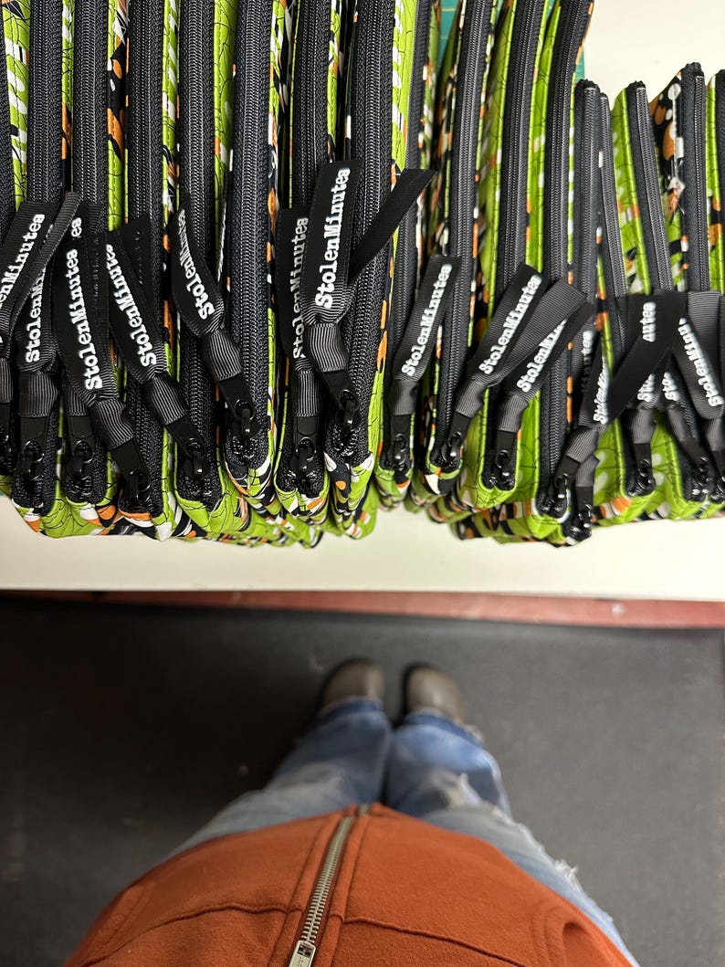 Puede incluir: Primer plano de varias bolsas con cremallera y cremalleras y tiradores negros. Las bolsas tienen una tela estampada verde y negra. Cada tirador de cremallera tiene el texto "StolenMinutes" impreso.